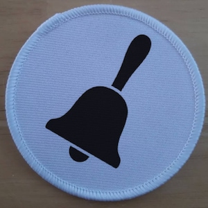 May include: A white round fabric patch with a black bell icon. The bell has a handle and a rounded bottom.