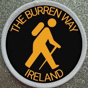 May include: A black and white circular patch with a white border. The patch features a yellow silhouette of a hiker with a backpack and walking stick. The text "THE BURREN WAY IRELAND" is written in yellow around the hiker.