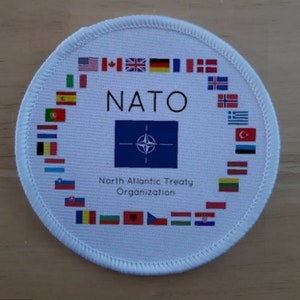May include: A white circular patch with the NATO logo and the text "NATO North Atlantic Treaty Organization". The patch is surrounded by a circle of flags representing the member countries of NATO.