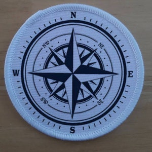 以下が含まれることがあります： 黒いコンパスデザインの白い丸い布のパッチ。コンパスは、北、南、東、西の主要な方向に伸びるポイントを持つ黒い星を特徴としています。コンパスには、北東、北西、南東、南西の中間方向も含まれています。