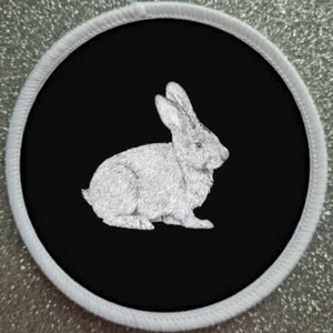 May include: A white rabbit is depicted in a black and white drawing on a black circular patch with a white border.