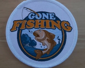 Może przedstawiać: Okrągła biała łatka z niebieskim i pomarańczowym wzorem. Wzór przedstawia rybę z napisem "GONE FISHING" pomarańczowymi literami.