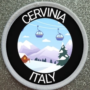 May include: A white and black round patch with the text "CERVINIA ITALY" in white. The patch features a cartoon illustration of a snowy mountain scene with a cabin, a pine tree, and three cable cars.