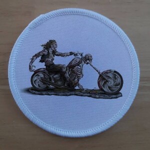 May include: A white round patch with a stitched edge features a black and white illustration of a skeleton riding a motorcycle. The skeleton is wearing a leather jacket and jeans. The motorcycle has a skull-shaped gas tank and a single headlight.