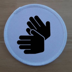 May include: White round fabric patch with a black silhouette of two hands clapping.