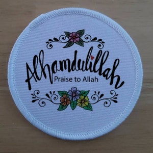 Puede incluir: Un parche redondo blanco con la frase "Alhamdulillah Praise to Allah" en escritura negra. El diseño incluye adornos florales en rosa, naranja, morado y amarillo, con un borde blanco cosido.