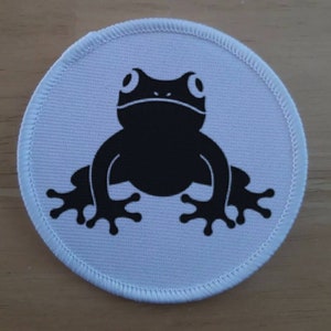 May include: A white round patch with a black silhouette of a frog. The frog has large eyes and is sitting on a white background.