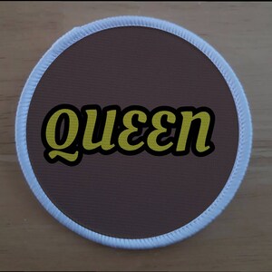 Könnte beinhalten: Ein runder brauner Patch mit weißem Rand. Das Wort "queen" ist in gelben Kursivbuchstaben geschrieben.