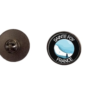 Op de afbeelding: Een ronde metalen speld met een afbeelding van een besneeuwd berglandschap met een kleine boom en skiliften. Het ontwerp is omringd door een zwarte rand met de witte tekst "SAINTE FOY" bovenaan en "FRANCE" onderaan.