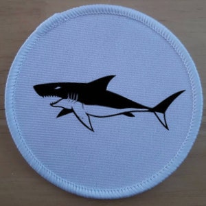 May include: A white round fabric patch with a black silhouette of a shark with a sharp fin and teeth.