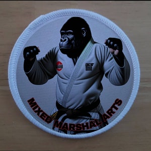 Puede incluir: Un parche circular blanco con una imagen en blanco y negro de un gorila que lleva un uniforme blanco de artes marciales con un cinturón negro. El texto "MIXED MARTIAL ARTS" está impreso en rojo alrededor del borde del parche.