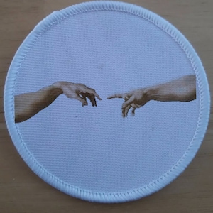 May include: A white round patch with a printed image of two hands reaching towards each other, reminiscent of Michelangelo's "The Creation of Adam" painting.