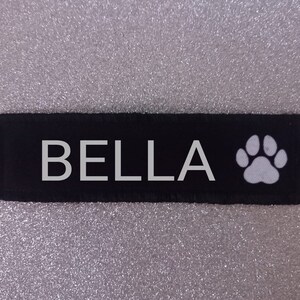 Puede incluir: Parche de tela negro con texto blanco que dice "BELLA" y una huella de pata blanca.