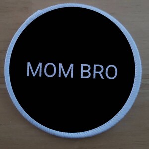 Op de afbeelding: Een ronde zwarte en witte patch met witte stiksels rond de rand. De tekst "MOM BRO" is in wit op een zwarte achtergrond gedrukt.
