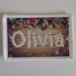 Puede incluir: Un parche de tela blanco con un diseño navideño. El parche presenta un fondo de madera con una guirnalda de ramas de pino, bayas rojas, piñas y un bastón de caramelo. El nombre "Olivia" está escrito en blanco sobre la madera.