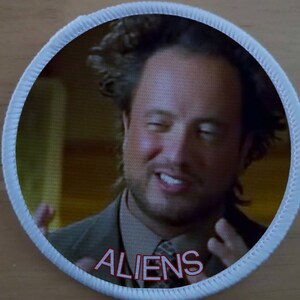 Può includere: Un cerotto rotondo bianco con un bordo rosso e la parola "ALIENS" stampata in rosso. Il cerotto presenta una foto di un uomo con un'espressione sorpresa.