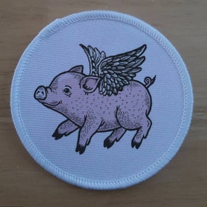 Puede incluir: Un parche redondo de color rosa claro con la ilustración de un cerdo con alas negras detalladas. El cerdo está representado en una pose de carrera, con una expresión sonriente. El parche tiene un borde blanco con detalles cosidos.