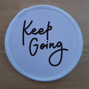 May include: A round, white patch with the words "Keep Going" in black script. The patch has a white stitched border. The text is centered on the patch.