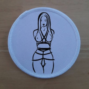 May include: A round, white patch with a black line drawing of a person in a bondage-style outfit. The illustration is centered on the patch, with a detailed outline of the figure's form and attire. The patch has a stitched border.