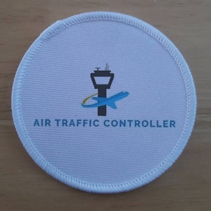 Può includere: Toppa rotonda bianca con un'immagine di un aereo blu e giallo e una torre di controllo. Il testo "AIR TRAFFIC CONTROLLER" è stampato in nero sotto l'immagine.