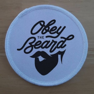 以下が含まれることがあります： 黒い文字と黒いひげのグラフィックが描かれた白い丸いパッチ。テキストは「Obey the Beard」です。