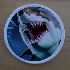 Könnte beinhalten: Ein weißer und grauer Hai-Patch mit offenem Maul, das scharfe Zähne zeigt. Der Patch ist rund und hat einen weißen Rand.