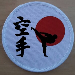 May include: A circular white embroidered patch featuring a black silhouette of a person performing a high kick. To the left of the figure is the black Japanese kanji for karate. A solid red circle is positioned behind the silhouette.