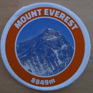 May include: A white and orange round patch with the text "MOUNT EVEREST" in a white circle on an orange background. The centre of the patch features a photo of Mount Everest with the text "8849m" below the image.