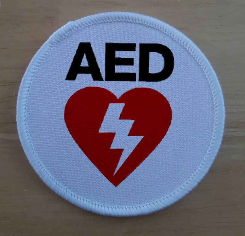 Może przedstawiać: Okrągła biała łatka z czerwonym sercem zawierającym błyskawicę i tekstem "AED" w kolorze czarnym.