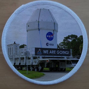 May include: A white spacecraft with the NASA and ESA logos is being transported on a flatbed trailer. The trailer has a banner that reads "We Are Going!" with the Artemis logo.