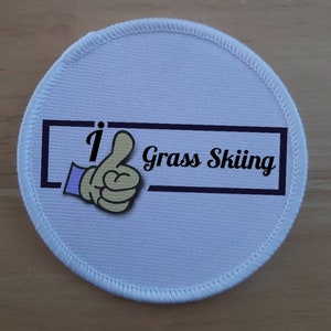 Op de afbeelding: Ronde witte patch met een zwarte rand en de tekst "I Grass Skiing" erin. Een cartoonhand met een duim omhoog staat links van de tekst.