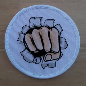 May include: White round patch with a stitched edge featuring a cartoon fist breaking through a ripped surface. The fist is brown and has a black outline.