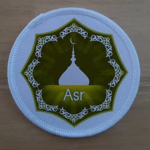 Puede incluir: Parche redondo de tela blanca con un diseño verde y blanco. El diseño presenta una mezquita estilizada con una luna creciente en la parte superior y la palabra "Asr" en letras blancas.
