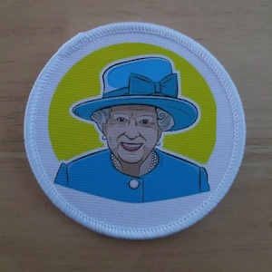 Könnte beinhalten: Ein runder weißer Aufnäher mit einem blauen und gelben Design. Der Aufnäher zeigt eine Comic-Abbildung von Königin Elisabeth II., die einen blauen Hut mit einer blauen Schleife und einen blauen Mantel trägt.