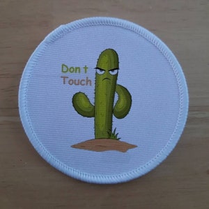 May include: A white round patch with a green cartoon cactus with a grumpy face and the text "Don't Touch" in green letters.