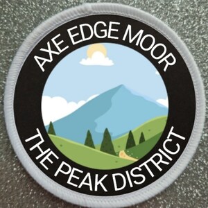 Op de afbeelding: Een witte en zwarte ronde patch met de tekst "Axe Edge Moor The Peak District" rond de rand. Het midden van de patch toont een cartoonillustratie van een bergketen met een blauwe lucht, witte wolken en een gele zon.