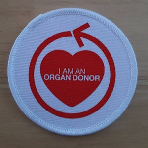 Könnte beinhalten: Runder weißer Aufnäher mit einem roten Herzen und einem roten Kreis mit einem Pfeil. Der Text im Herzen lautet "Ich bin ein Organspender".