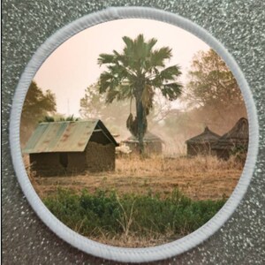 May include: A circular image of a rural African village with a tall palm tree in the center. The village is made up of several small, mud-brick houses with thatched roofs. The houses are surrounded by dry, brown grass and a few trees.
