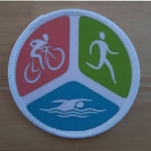 May include: A white, round patch with a tri-color design. The top left section is red with a white silhouette of a person riding a bicycle. The top right section is green with a white silhouette of a person running. The bottom section is blue with a white silhouette of a person swimming.