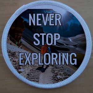May include: White round patch with a blue and white mountain scene and the text "NEVER STOP EXPLORING" in black.