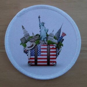 May include: A white round patch featuring a colourful image of an American flag suitcase with iconic landmarks of New York City, including the Statue of Liberty, the Empire State Building, and the Chrysler Building.