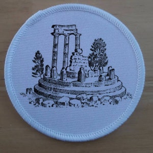 May include: A white fabric patch featuring a black and white illustration of an ancient Greek temple ruin. The temple is surrounded by trees and rocks.