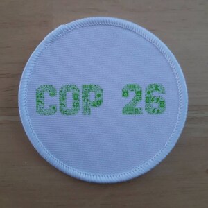 May include: A white, circular patch with the text "COP 26" in green. The text is composed of small, green icons related to environmental sustainability. The patch has a white border with a stitched edge.