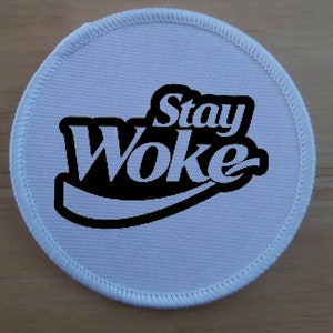 May include: White round fabric patch with black stitching around the edge. The patch has a black and white graphic that says "Stay Woke" in a stylized font.
