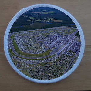 Peut inclure: Vue aérienne d'un grand circuit ovale entouré d'un grand parking. Le circuit est entouré d'arbres et d'une petite ville.