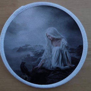 Pode incluir: Uma mulher vestida de branco com cabelo branco comprido senta-se em uma costa rochosa, olhando para baixo para um corvo preto. A cena é nebulosa e etérea, com um fundo azul suave.