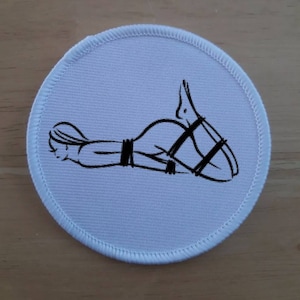 May include: A white round patch with a black line drawing of a person tied up with rope.