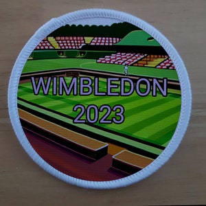 Könnte beinhalten: Ein weißer runder Patch mit einem grünen Tennisplatz, roten und weißen Tribünen und dem Text "WIMBLEDON 2023" in Lila.