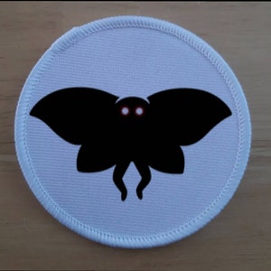 Op de afbeelding: Een ronde witte patch met een zwart silhouet van een mot met rode ogen.