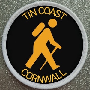 Pode incluir: Um remendo redondo branco e preto com o texto "TIN COAST CORNWALL" em amarelo. Uma silhueta amarela de uma pessoa com uma mochila e um bastão de caminhada está no centro.